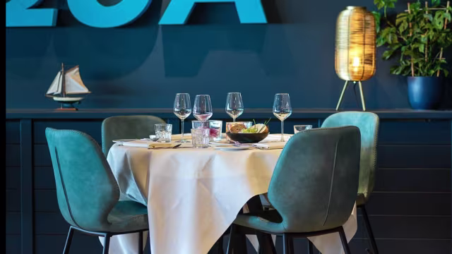 Moderne eetruimte met een strak wit tafelkleed, wijn glazen, een klein kommetje met olijven en een opvallende blauwe muur met een "20A" bord, wat bijdraagt aan de uitnodigende sfeer.