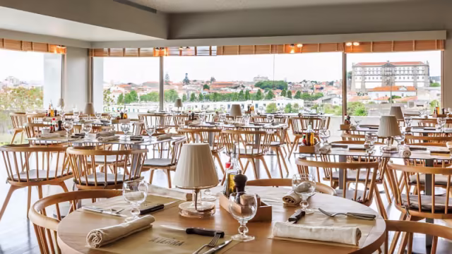 Interior do restaurante com um layout espaçoso, mesas e cadeiras de madeira, criando uma atmosfera acolhedora, iluminada por janelas grandes que oferecem vistas panorâmicas. A decoração minimalista e elegante realça o ambiente.