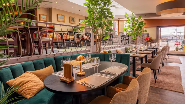 Moderne restaurantinterieur met een ruime indeling, comfortabele groene banken en donkere houten tafels. De decoratie omvat potplanten en grote ramen die natuurlijk licht binnenlaten, wat zorgt voor een gezellige en elegante sfeer.