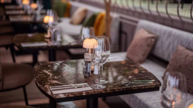 Stijlvol restaurantinterieur met elegant gedekte tafels met donkere marmeren bladen, gepolijst glaswerk en net gerangschikte bestek. Zachte, sfeervolle verlichting van kleine lampen creëert een warme en uitnodigende sfeer, terwijl comfortabele banken met kleurrijke kussens de esthetiek van de ruimte versterken.