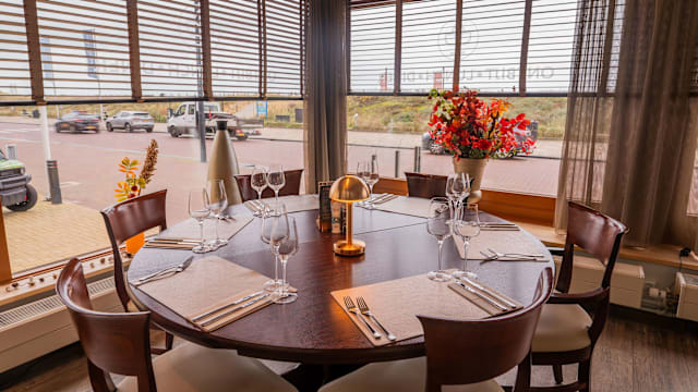 Een elegant gedekte ronde houten tafel met bestek en glazen, omringd door grote ramen die natuurlijk licht binnenlaten. Een vaas met kleurrijke bloemen voegt warmte en frisheid toe aan de sfeer.