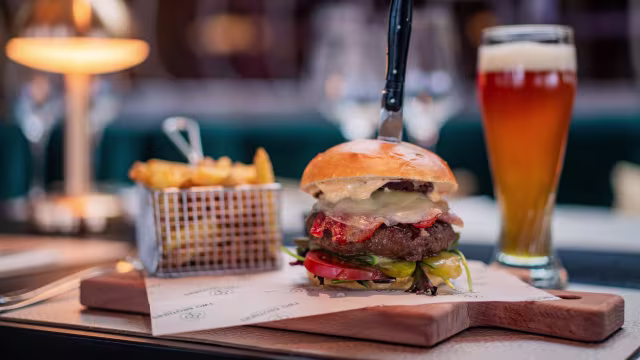 Een sappige burger met een dikke beefburger, gesmolten kaas en verse toppings zoals sla, tomaat en augurken, geserveerd in een zacht, geroosterd broodje. Vergezeld van goudgele, knapperige frietjes in een klein draadmandje en een verfrissend amberbier in een hoog glas.