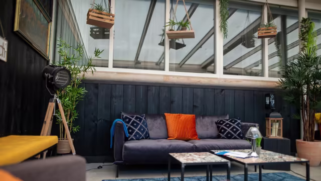 Gezellige restaurantinterieur met een moderne esthetiek, inclusief een diepe blauwe bank met decoratieve kussens, kleine tafels met verschillende texturen en hangplanten die de uitnodigende sfeer versterken. Natuurlijke lichtinval door grote ramen creëert een warme ambiance.