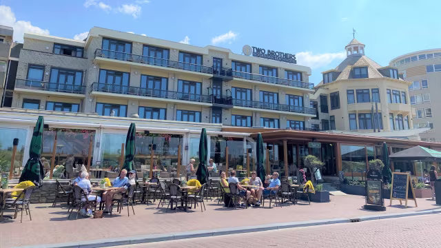 Restaurantgevel met een modern ontwerp, grote glazen ramen en een buitenzitgedeelte met tafels en stoelen, waar gasten in een ontspannen sfeer genieten van hun maaltijden.