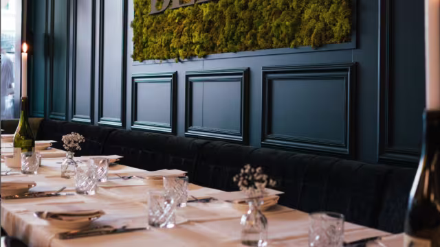 Stijlvol restaurantinterieur met een elegante wand van groen mos, een gezellige donkergekleurde bank en netjes gedekte tafels met witte tafelkleden en eenvoudige bloemstukken.