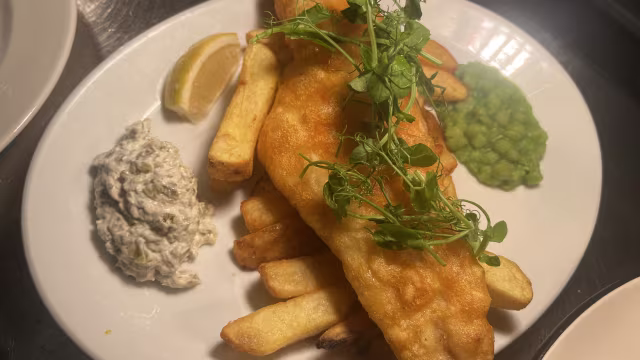 Golden-battered fish fillet served with thick-cut fries, garnished with fresh herbs, lemon wedges, mushy peas, and creamy tartar sauce.