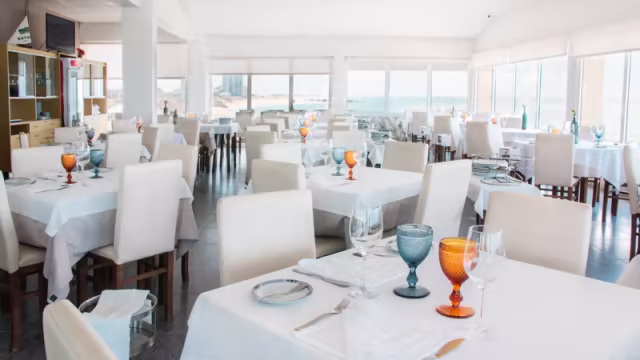 Interior do restaurante com uma atmosfera luminosa e arejada, grandes janelas, mesas elegantemente postas com toalhas brancas e talheres organizados, cadeiras de cores claras e decoração minimalista, criando um ambiente relaxante.