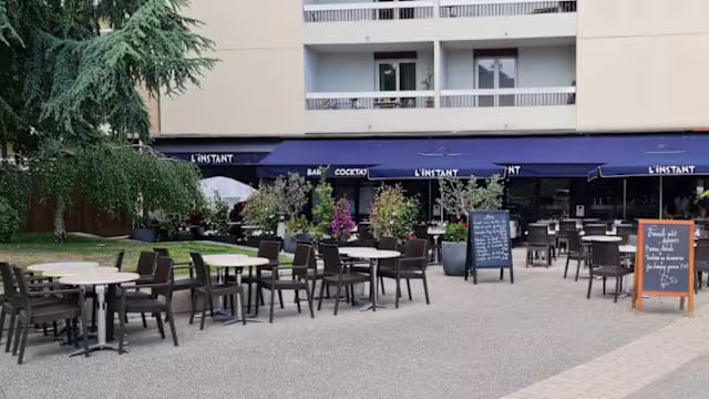 Espace de restauration en plein air avec des tables modernes et des chaises sombres, agrémenté de plantes en pot et d'un tableau noir affichant le menu.