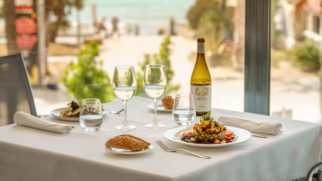 En la mesa se presentan tres platos: una ensalada colorida con verduras frescas, tomates cherry y pepino; un plato de pasta con verduras y salsa rica, decorado con hierbas; y un pequeño filete de pescado a la parrilla con una rodaja de limón. Además, hay tres copas de vino blanco y un panecillo dorado.