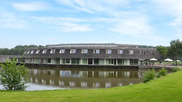 Modern restaurant met een unieke ronde vorm, gelegen aan een rustige waterpartij, omgeven door een goed onderhouden gazon en buitenzitplaatsen met parasols.