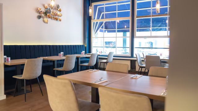 Het interieur van het moderne restaurant is schoon en uitnodigend, met houten tafels en lichte stoelen voor een informele eetervaring. Een blauwe beklede bank langs de muur voegt comfort toe. De minimalistische decoratie, met een wandstuk van gouden en zilveren elementen, straalt subtiele elegantie uit. Grote ramen zorgen voor veel natuurlijk licht, wat de sfeer versterkt. De indeling is ruim, geschikt voor zowel kleine als grotere groepen.