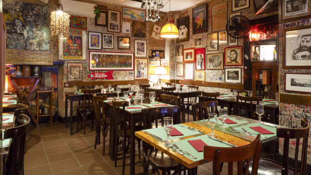 Interno di un ristorante accogliente con opere d'arte e fotografie, tavoli e sedie in legno, illuminazione soffusa e un'atmosfera intima.