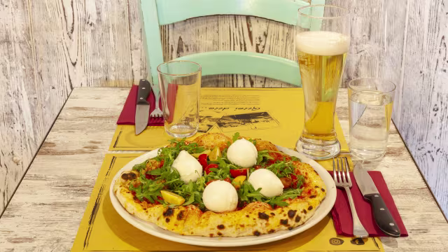 Pizza rotonda con mozzarella fresca, rucola, peperoni rossi e pomodorini, servita con birra chiara.
