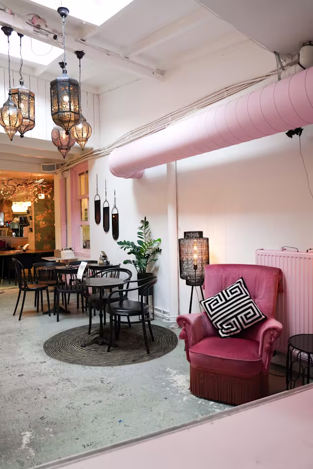 Een charmant restaurantinterieur met een mix van moderne en vintage decoratie, verlicht door decoratieve lantaarns. Een roze, gebogen luchtkanaal voegt een speelse toets toe. Een pluche, roze fauteuil met een zwart-wit gestreepte kussen en een rond tapijt creëren een gezellige zithoek met zwarte tafels en stoelen. Potplanten zorgen voor een vleugje groen en versterken de uitnodigende sfeer.