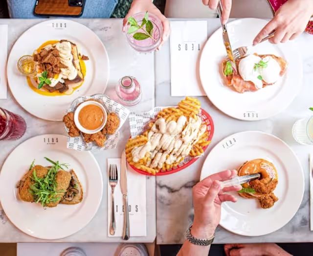 Een levendige eettafel met verschillende gerechten: knapperige gefrituurde kip met een klein schaaltje dipsaus en gouden frietjes, mooi opgemaakte eggs benedict met gepocheerde eieren op gerookte zalm en verse kruiden, en een bord met hartig vlees, mogelijk pulled pork, met een rijke saus en romige aardappelpuree. De tafel is versierd met kleurrijke dranken en een stijlvolle marmeren oppervlakte.