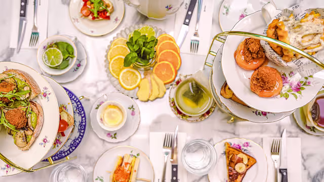 Elegante afternoon tea met een etagère vol gebakjes en hartige hapjes. De bovenste laag bevat delicate gebakjes met room en fruit, de middelste laag heeft kleine sandwiches met verse groenten, en de onderste laag bestaat uit scones, geserveerd met clotted cream en jam. Om de etagère liggen kleurrijke salades en fruitschijven, waaronder sinaasappels en citroenen.