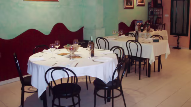 Interior de un restaurante acogedor con mesas redondas, manteles blancos y paredes turquesa con acentos rojos, creando un ambiente cálido y espacioso.