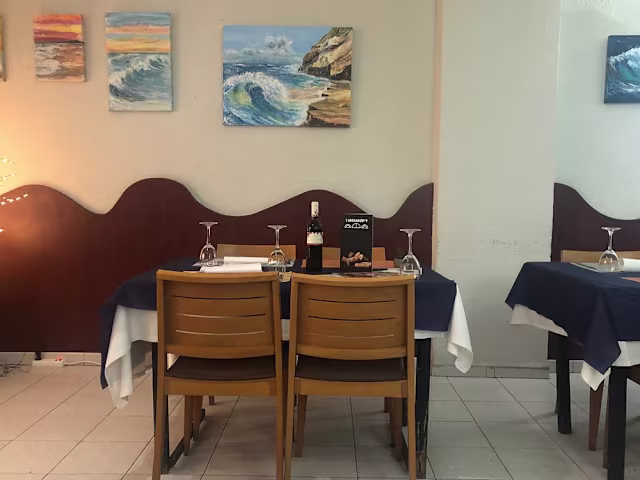 Interior de restaurante acogedor con mesas y sillas de madera, manteles azules y pinturas coloridas de olas del océano en las paredes, creando un ambiente cálido y acogedor.