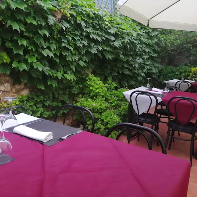 Área de comedor al aire libre con mesas y sillas de estilo bistró, decoradas con manteles rojos y rodeadas de vegetación exuberante.