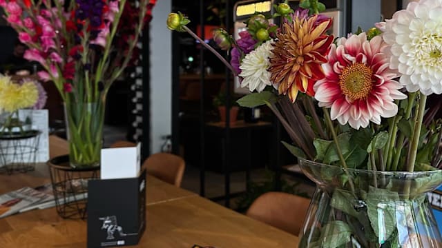 Gezellig restaurantinterieur met grote, kleurrijke bloemstukken van dahlia's en gladiolen in diepe roodtinten, heldere geeltinten en zachte witten, geplaatst in glazen vazen en draadmanden.