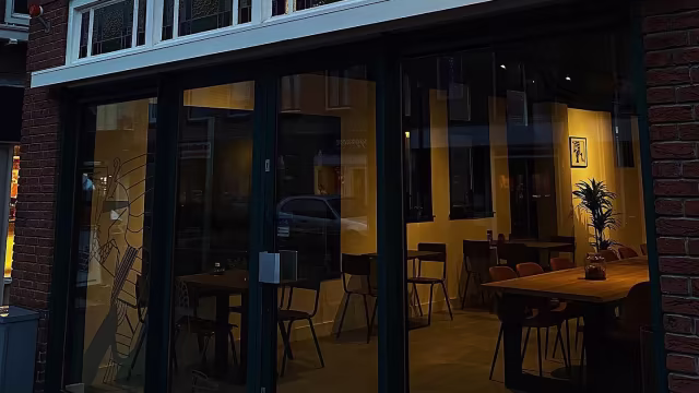 Het exterieur van een restaurant met grote glazen ramen die een warm en uitnodigend interieur onthullen, verlicht met zachte verlichting en een moderne inrichting. Binnen zijn er houten tafels en stoelen, wat bijdraagt aan een gezellige sfeer.