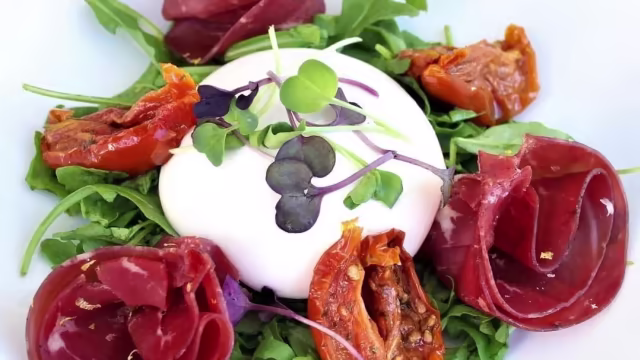 Ensalada fresca con burrata cremosa, rúcula, tomates secos y finas lonchas de jamón curado, decorada con microgreens.