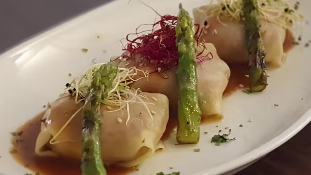 Tres dumplings envueltos en masa translúcida, acompañados de espárragos a la parrilla, salsa sabrosa, brotes de colores y semillas de sésamo.