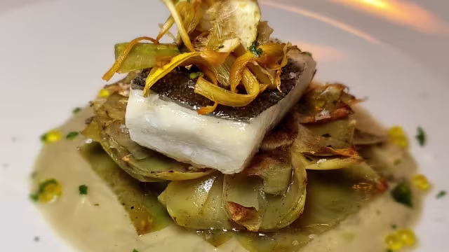Pescado blanco perfectamente cocido sobre corazones de alcachofa, acompañado de cebollas fritas crujientes, verduras en rodajas y una suave salsa cremosa con hierbas.