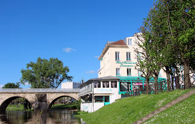 Restaurant charmant au bord de l'eau, avec une façade claire, de grandes fenêtres et une terrasse extérieure couverte ornée de parasols colorés, offrant une ambiance accueillante.