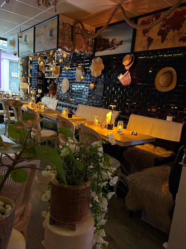 Intérieur de restaurant charmant alliant décor rustique et côtier, avec des murs en carreaux noirs, des chapeaux décoratifs et un éclairage chaleureux, créant une atmosphère accueillante. Tables en bois et sièges confortables, agrémentés de plantes en pot, pour une expérience de repas détendue et conviviale.