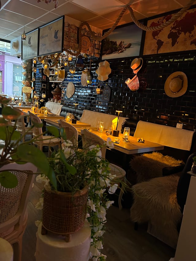 Intérieur de restaurant charmant avec un mélange de décor rustique et côtier, des murs ornés de chapeaux et d'objets décoratifs, et un éclairage doux provenant de bougies. Mobilier confortable avec des chaises rembourrées et des tables en bois, agrémenté de plantes en pot pour une ambiance accueillante.