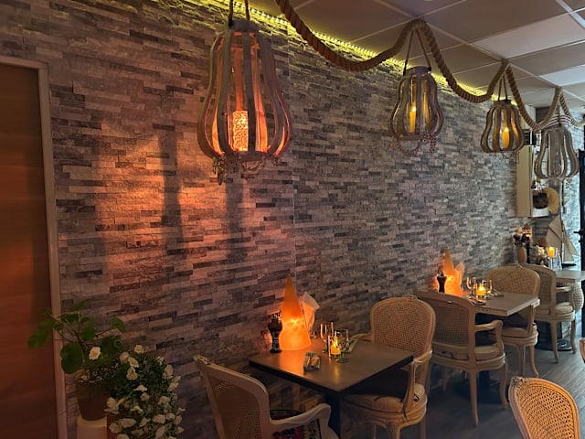 Intérieur de restaurant chaleureux avec un mur en pierre texturé, éclairé par une lumière douce. Tables en bois et chaises en osier créent une ambiance détendue, agrémentée de luminaires suspendus uniques et de petites bougies sur les tables. Des plantes en pot et des éléments décoratifs renforcent l'atmosphère accueillante.