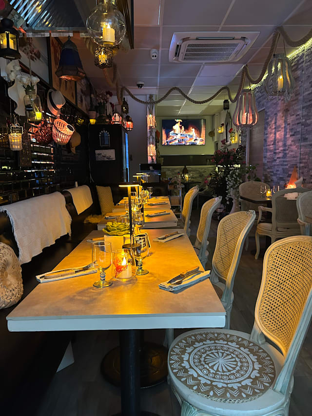 Intérieur de restaurant chaleureux avec une grande table dressée, éclairée par des lumières décoratives et ornée d'éléments artistiques. Ambiance accueillante avec des bougies et des sièges confortables.