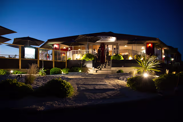 Extérieur d'un restaurant moderne à la tombée de la nuit, avec un éclairage doux, une entrée accueillante, des plantes décoratives et des parasols pour les espaces extérieurs.