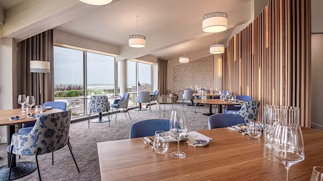 Intérieur de restaurant moderne et spacieux, avec de grandes fenêtres laissant entrer la lumière naturelle, des éléments en bois et des textiles doux, créant une ambiance contemporaine et accueillante.