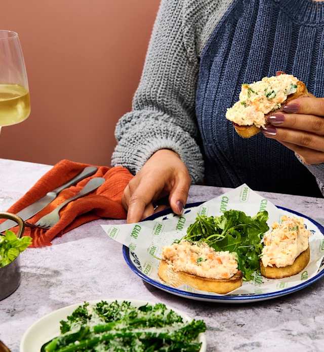 Toasted bread topped with a creamy seafood spread, likely crab or shrimp, garnished with fresh herbs and accompanied by a side of leafy greens.