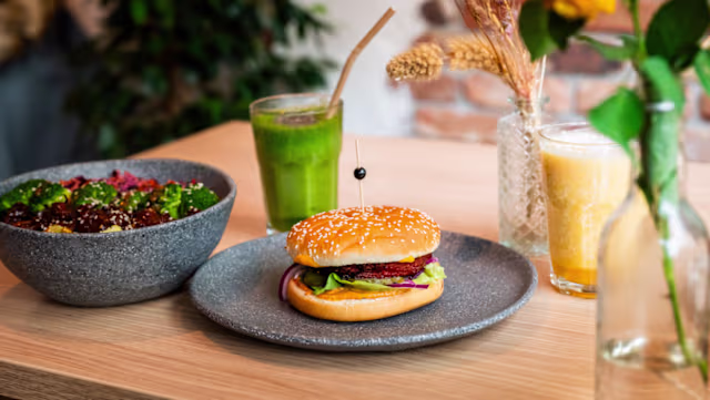 Sandwich met sesamzaadjes, gevuld met verse groenten zoals sla, tomaat en augurken, en een gegrilde burger. Geserveerd met een kleurrijke salade en twee verfrissende dranken: een groene smoothie en een romige drank.