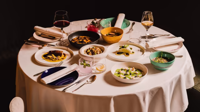 Tavolo elegantemente apparecchiato con piatti di carne brasata, pasta cremosa, frutti di mare, insalata mista e torta al cioccolato, accompagnati da vini rossi e bianchi.