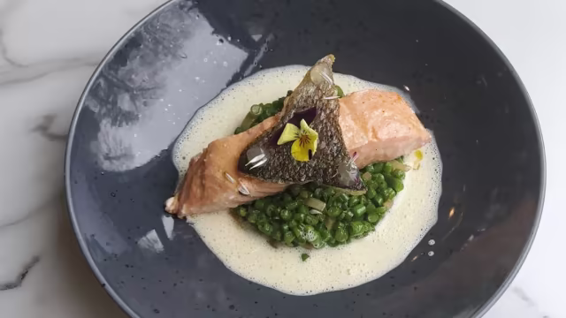 Plat de saumon tendre légèrement poêlé, avec une peau croustillante, servi sur un lit de petits pois finement hachés et accompagné d'une sauce crémeuse délicate, le tout agrémenté d'une fleur comestible.
