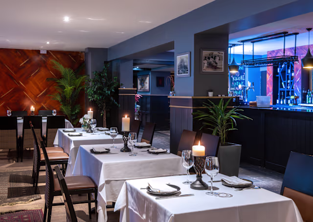 A modern restaurant interior with elegantly set tables, soft lighting, potted plants, framed artwork, and a sleek bar area.
