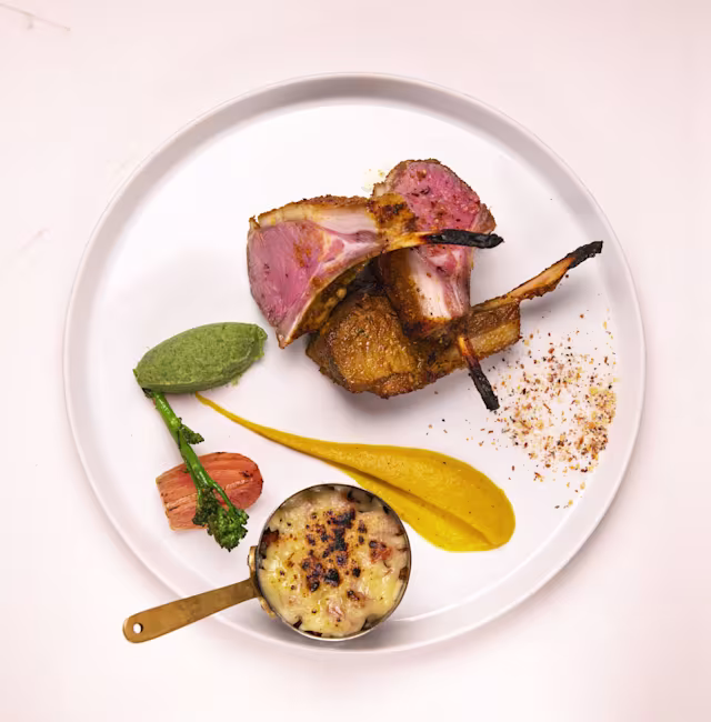 Chargrilled lamb chops with a golden crust, served with a bright orange sauce, creamy potato gratin, a green herb puree, and a pink pickled garnish.
