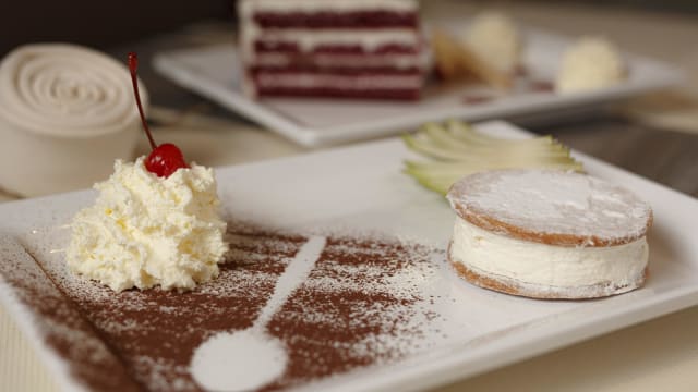 Dessertpresentatie met een delicate sandwichkoek gevuld met romige vulling, bestrooid met poedersuiker, geserveerd met een toef slagroom en een rode kers, naast een plak gelaagde taart.