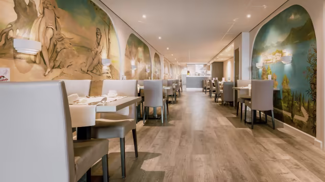 Het interieur van het restaurant heeft een moderne en uitnodigende uitstraling, met lichte muren versierd met grote, artistieke muurschilderingen van serene landschappen en klassieke figuren. De vloer is van warm hout, wat bijdraagt aan de gezellige sfeer. Tafels zijn netjes gedekt met witte tafelkleden en eenvoudige servies, terwijl comfortabele, lichtgekleurde stoelen zorgen voor extra comfort. Zachte wandverlichting creëert een warme ambiance, waardoor de ruimte uitnodigend en elegant aanvoelt. De indeling suggereert een ruime eetzaal, ideaal voor zowel intieme maaltijden als grotere bijeenkomsten.