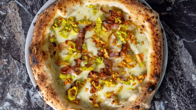 Pizza appena sfornata con crosta dorata e leggermente bruciacchiata, con salsa bianca cremosa, pancetta croccante e porri dolci.