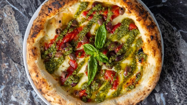 Pizza appena sfornata con crosta dorata e leggermente bruciacchiata, con salsa bianca cremosa, pesto verde, peperoni arrostiti e olive nere.