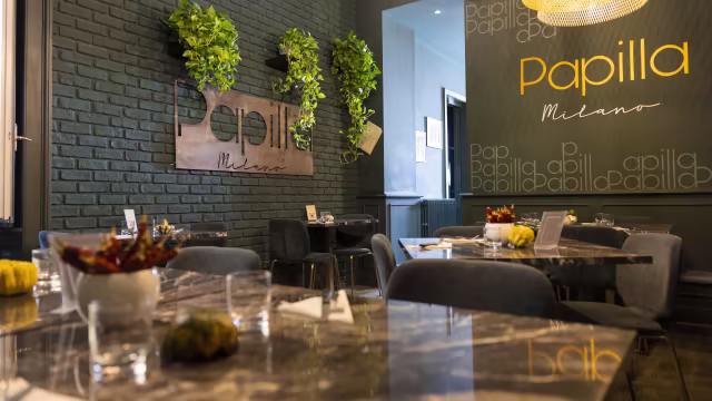 Interni moderni e accoglienti con pareti verde scuro e piante, tavoli in marmo lucido e sedie grigie eleganti, creando un'atmosfera contemporanea e rilassante.