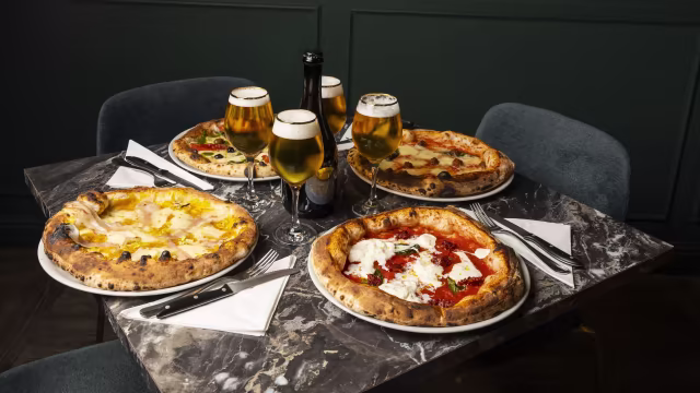 Selezione di quattro pizze con crosta spessa e bruciata, preparate in forno a legna: pizza con formaggio dorato e olive nere, pizza Margherita con salsa di pomodoro e basilico, pizza con base di salsa bianca, prosciutto e rucola, e pizza con funghi e olio d'oliva.