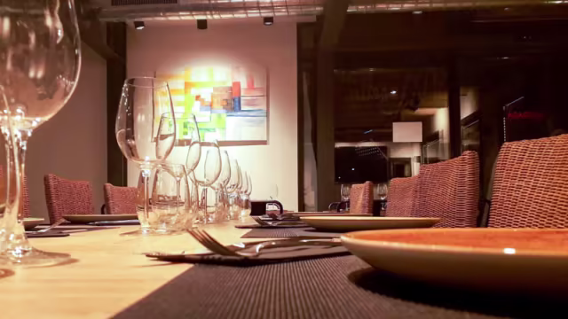 Interior de restaurante con una mesa de madera larga, decorada con cristalería elegante y arte abstracto en la pared, creando un ambiente moderno y acogedor.