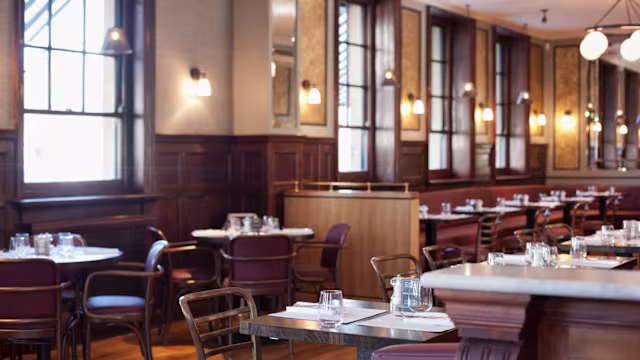 Cozy restaurant interior with wooden paneling, large windows, white tablecloths, and soft lighting, featuring a mix of wooden chairs and plush banquettes for a comfortable dining experience.