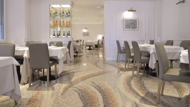Interni moderni ed eleganti con toni neutri, pavimento decorato, tavoli con tovaglie bianche e sedie imbottite. Atmosfera luminosa e accogliente con illuminazione sottile e opere d'arte.